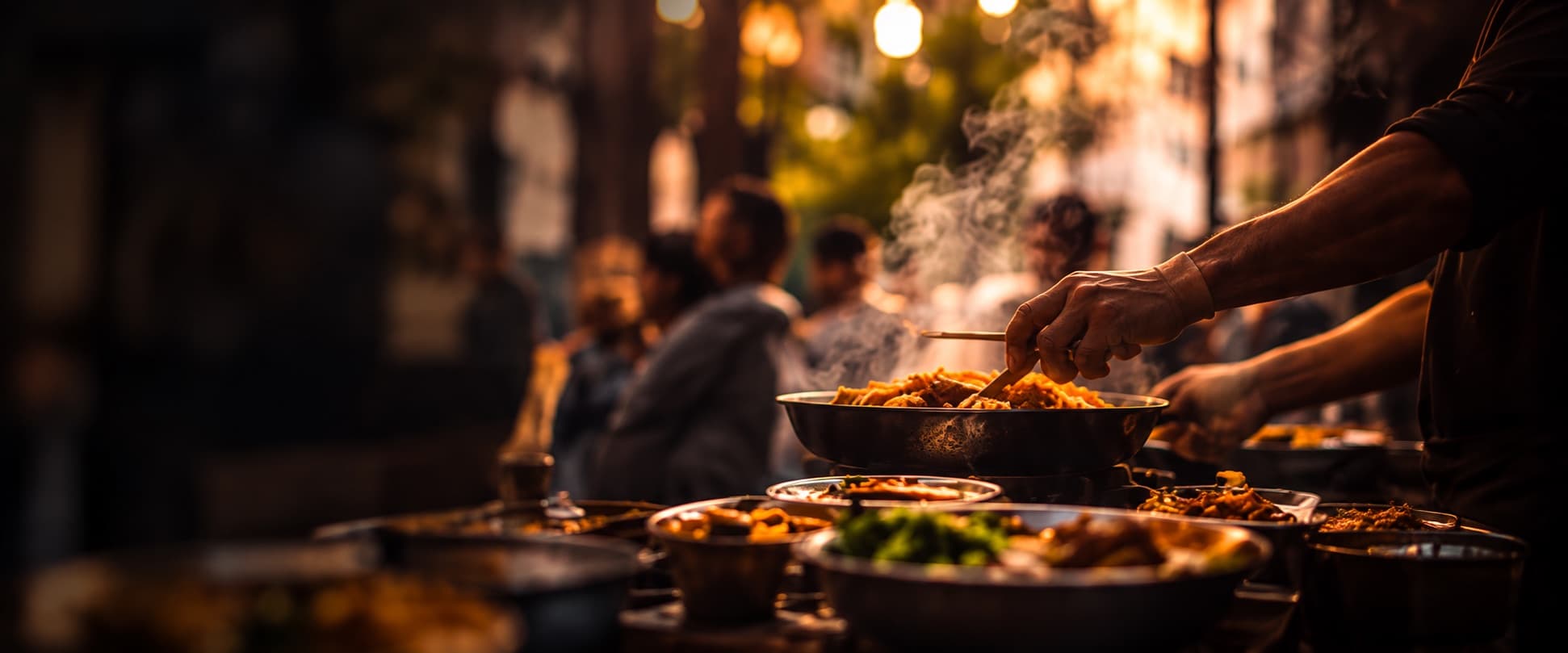A vendor's hands serving hot food from a pan at a night street food gathering, steam rising